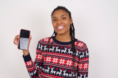 Beyaz duvara karşı Noel süveteri giyen gülümseyen Afro-Amerikan kadın boş telefon ekranı gösteriyor. Reklam ve iletişim konsepti.