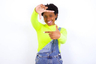 Kısa saçlı, neşeli ifadeli, beyaz duvara karşı kot pantolon giyen pozitif bir Afro-Amerikalı kadın. İyi bir ruh hali var. Kameraya doğru hareketler yapıyor..