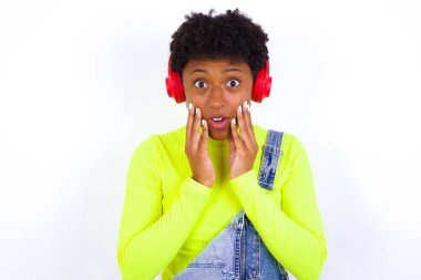 Şok olmuş, kısa saçlı, beyaz pantolonlu, kameradan korkan, ağzı açık kulaklık takan, kısa saçlı, Afro-Amerikan bir kadın.