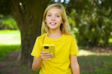 Açık havada duran sarı tişörtlü beyaz kız cep telefonunu elinde tutuyor ve olumlu haberler veriyor, modern cep telefonu kullanıyor.