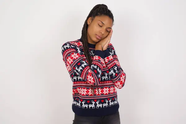 Relax and sleep time. Tired African American woman wearing Christmas sweater against white wall with closed eyes leaning on palms making sleeping gesture.