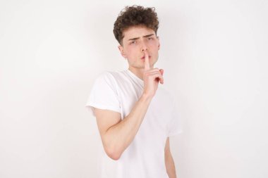 Şaşırmış genç adam sessiz bir jest yapıyor, parmaklarını dudaklarının üstünde tutuyor ve kameraya gizemli bakıyor.