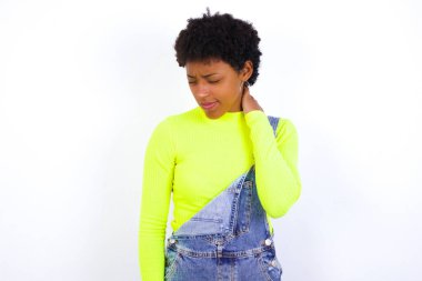 Beyaz duvara karşı kot pantolon giyen, sırtı ve boynu ağrıyan, eliyle boynuna dokunan kısa saçlı, genç Afrikalı bir kadın..