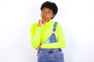 Yüz ifadeleri ve duygular. Kısa saçlı, düşünceli, genç Afrikalı Amerikalı kadın. Kot pantolon giyip beyaz duvara yaslanmış. Başının altında el ele tutuşmuş..