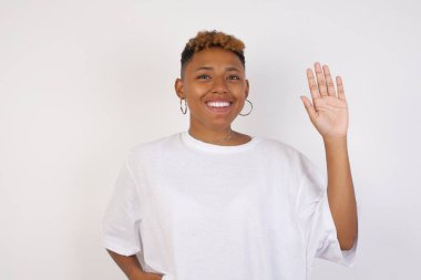 Genç, güzel, Afro-Amerikalı bir kadın beyaz tişörtlü, izole edilmiş arka planda duruyor. Mutlu ve güler yüzlü, dostça bir jest yapıyor.