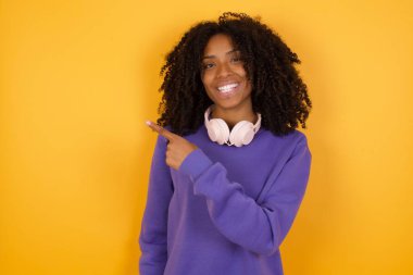 Sarı arka planda kulaklıkları olan, boş bir yeri işaret eden Afro-Amerikan genç kadının portresi.