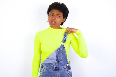 Beyaz duvara karşı kot pantolon giyen genç Afrikalı Amerikalı kadın stresli, endişeli, yorgun ve hayal kırıklığına uğramış, gömleğini çekiştiren, sorunlardan bıkmış görünüyor.