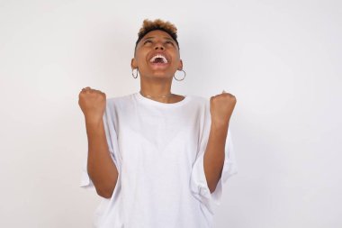 Beyaz tişört giyen güzel Afro-Amerikan kadın portresi heyecanla bakıyor, ellerini havada tutuyor, beklenmedik bir şey fark ediyor, içeride poz veriyor. Güzel bir kadın ani haberlere tepki verir..