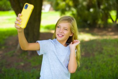 Beyaz tişörtlü mutlu beyaz kız çocuğu parkta dikiliyor cep telefonu kullanıyor ve selfie çekiyor.