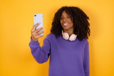 Sarı arka planda kulaklıklı Afro-Amerikan kadının portresi akıllı telefonlu selfie çekiyor.