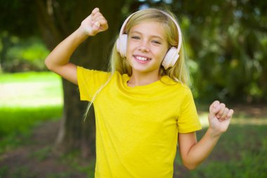 Kafkasyalı, sarı tişörtlü, dişlek bir şekilde dışarıda dikilen, kollarını kaldıran, dans eden, ritimli, müzik dinleyen kulaklıkla müzik çalan küçük kız.
