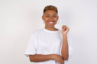 Beyaz tişört giyen güzel, Afro-Amerikalı bir kadının gri duvarın karşısında eli yüzüne yakın duruşu. İyi haberleri dinledikten sonra kameraya mutlu bir ifadeyle bakmak. Kendine güvenen kız..