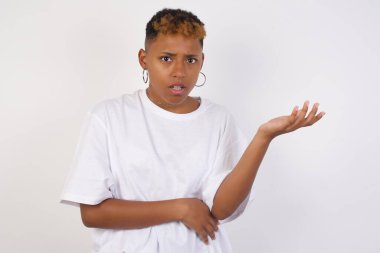 What the hell are you talking about, nonsense. Studio shot of frustrated young African American woman wearing white t-shirt gesturing with raised palm, frowning, being displeased and confused with dumb question over gray wall.