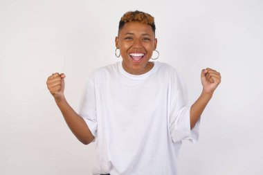 Beyaz tişört giyen, sıradan tişörtlü, kollarını kaldıran, yumruklarını sıkan, neşe ve heyecanla bağıran güçlü ve kararlı genç Afrikalı Amerikalı kadının portresini kaldır. Zafer, başarı ve başarı kavramı.