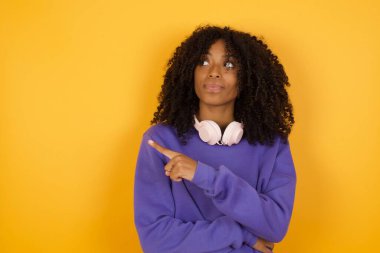 Sarı arka planda kulaklıkları olan, boş bir yeri işaret eden Afro-Amerikan genç kadının portresi.