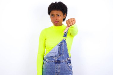 Beyaz duvarlara karşı kot pantolon giyen hoşnutsuz genç Afrikalı Amerikalı kadın onaylamadığını gösteriyor, başparmağını aşağıda tutuyor, hoşnutsuzluğunu ifade ediyor, hoşnutsuzluğunu ifade ediyor. Olumsuz duygular.