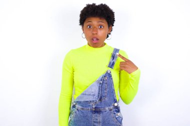 Beyaz duvara karşı kot pantolon giyen kısa saçlı genç Afrikalı Amerikalı kadın sersemlemiş bir şekilde kendini parmağıyla işaret ederek 