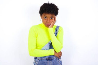 Beyaz duvara karşı kot pantolon giyen üzgün, yalnız, genç Afrikalı Amerikalı kadın yanağına dokunuyor alt dudağını ısırıyor ve hoşnutsuzca bakıyor. Kötü duygular