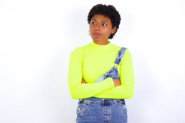 Beyaz duvara karşı kot pantolon giyen düşünceli, düşünceli, genç Afrikalı Amerikalı kadın kollarını kavuşturmuş, düşünceli bir ifadeyle ne yapacağını düşünüyor.