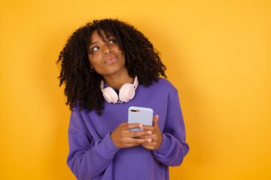 Sarı arka planda kulaklıklı Afro-Amerikan kadının akıllı telefon kullanarak portresi.