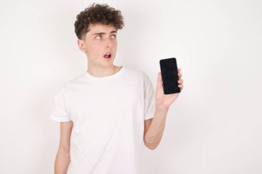 Beyaz arka plandaki yakışıklı genç adam yeni cep telefonunu tutuyor ve gizemli görünüyor. Modern cep telefonunun boş görüntülerini gösteriyor.