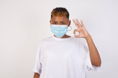 Beyaz tişört giyen çekici, Afro-Amerikalı genç bir kadının, olumlu bir ifade ve iyimser bir ifade sergilemesine sevindim. Beyaz duvara yaslanarak.