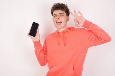 Beyaz arka plan üzerinde heyecanlı genç bir adam akıllı telefon boş ekranı, göz kırpması ve el işareti yapıyor. Reklam kavramı.