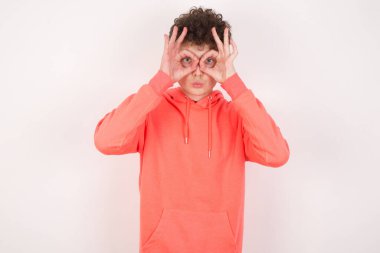 Beyaz arka planda neşeli, heyecanlı genç bir adam. Tamam işareti yaparken iki gözünün üstünde, gözlük takıyormuş gibi yapıyor..