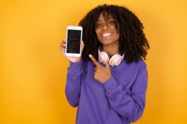 Sarı arka planda kulaklıklı Afro-Amerikan kadının akıllı telefonu gösteren portresi.