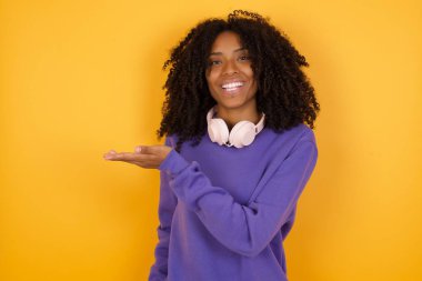 Sarı arka planda kulaklıkları olan, boş bir yeri işaret eden Afro-Amerikan genç kadının portresi.