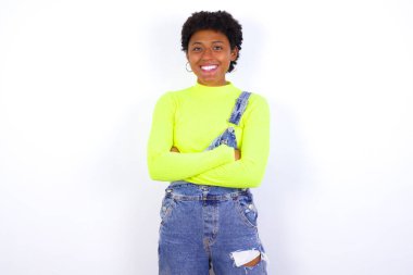 Beyaz duvara karşı kot pantolon giyen genç Afrikalı Amerikalı kadın gülümsüyor ve kameraya kendinden emin bir şekilde kollarını kavuşturuyordu. Pozitif ve kendinden emin kişi.