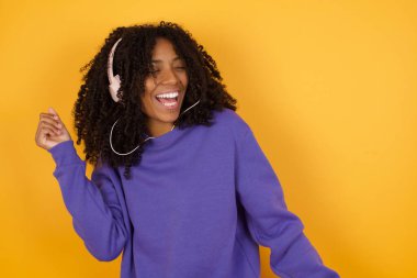 Sarı arka planda müzik dinleyen kulaklıklı Afro-Amerikan kadının portresi.