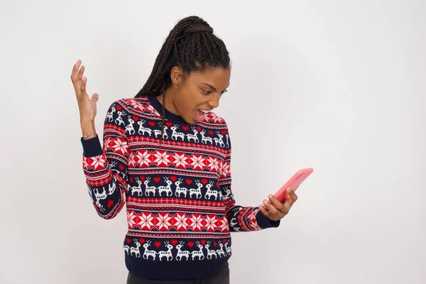Photo of outraged annoyed African American woman wearing Christmas sweater against white wall holds cell phone, makes call, argues with colleague,  expresses negative emotions. People and anger.
