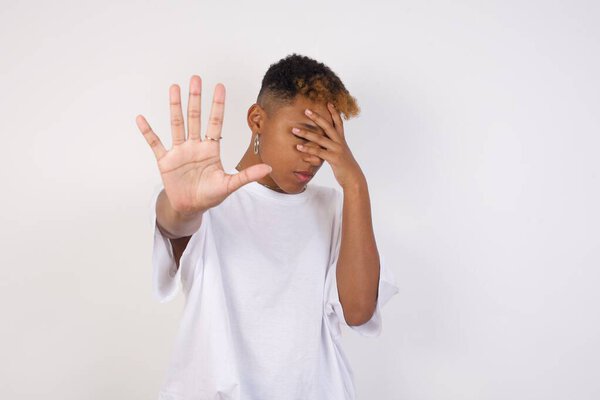 Young black woman covers eyes with palm and doing stop gesture, tries to hide from everybody. Don't look at me, I don't want to see, feels ashamed or scared.