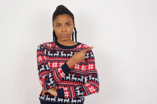 身穿圣诞毛衣 靠着白墙的非洲裔美国女人对着摄像机笑着 指指点点着自己的手指 表现出一些有趣而令人兴奋的东西 — 图库照片