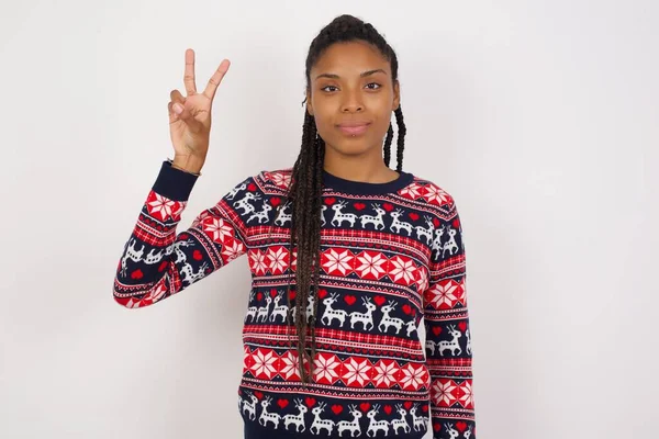 アフリカ系アメリカ人女性が白い壁にクリスマスセーターを着て 自信と幸せを笑いながら指ナンバー2を指で指で指で指を指さす — ストック写真