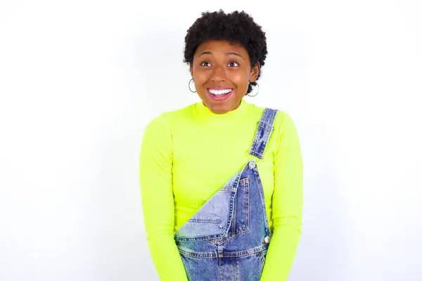 Překvapená Afroameričanka Krátkými Vlasy Džínové Kombinéze Opřenými Bílou Stěnu Pokrčí — Stock fotografie