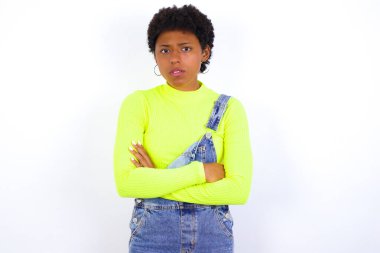 Kısa saçlı Amerikalı bir kadın, beyaz duvara karşı kot pantolon giyiyor, yüzünü hoşnutsuzca çatıyor, kollarını kavuşturup, bir açıklama bekliyor..