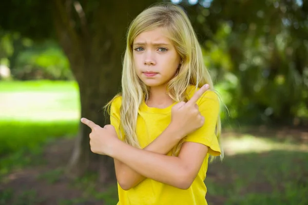 严重的高加索小女孩穿着黄色的T恤站在室外交叉双手 不同侧面的点在两件物品之间踌躇 硬性决策概念 — 图库照片