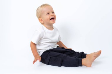 Beyaz tişört ve siyah pantolon giyen sarışın çocuk yerde oturuyor ve beyaz stüdyo arka planında poz veriyor.