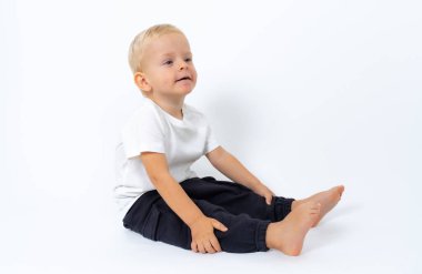 Beyaz tişört ve siyah pantolon giyen sarışın çocuk yerde oturuyor ve beyaz stüdyo arka planında poz veriyor.