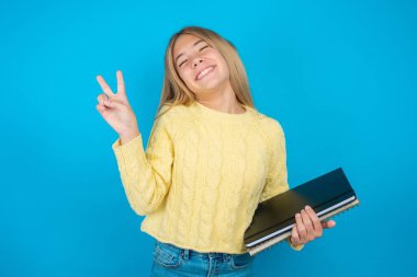 Mavi arka planda sarı süveter giyen, gülümseyen ve kameraya bakıp zafer işareti yapan güzel bir kız çocuğu. İki numara..