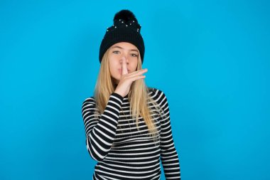 Güzel bir kız, siyah şapkalı ve mavi arka planda çizgili balıkçı yaka giyen sessiz hareketler yapan, parmaklarını dudaklarının üzerinde tutan. Sessizlik ve gizli konsept.
