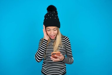 Güzel bir kız, örülmüş siyah şapka ve çizgili balıkçı yaka giyen mavi arka planda elinde cep telefonuyla yeni uygulama 5G kullanarak SMS okuyor.