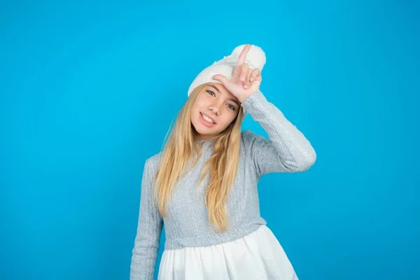 ホワイトニットの帽子と青のセーターを着用した美しい子供の少女は 失われたジェスチャーを嘲笑し侮辱している額に指を持つ人々を楽しみます — ストック写真