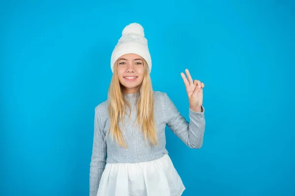 Красивая Девочка Белой Вязаной Шляпе Голубом Свитере Показывая Показывая Пальцами — стоковое фото