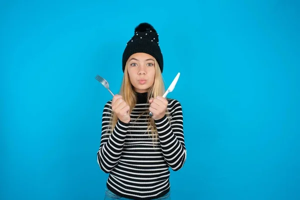 hungry beautiful kid girl wearing knitted black hat and striped turtleneck over blue background holding in hand fork knife want tasty yummy pizza pie