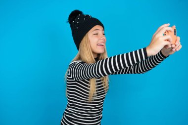 Güzel bir kız çocuğu örülmüş siyah şapka ve mavi arka plan üzerine çizgili balıkçı yaka giyip sosyal medyada yayınlamak için selfie çekiyor ya da arkadaşlarıyla video görüşmesi yapıyor..