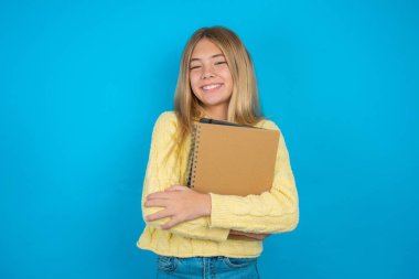 Kendine güveni tam, sakin, mavi arka planda sarı süveter giyen, kolları bağlı güzel bir kız çocuğu. Profesyonel titreşimlerin kendine güvenen pozlarda durduğunu gösterir.