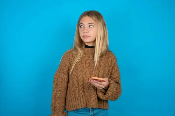 Blondes Mädchen Mit Braunem Strickpullover Vor Blauem Hintergrund Hält Mobiltelefone — Stockfoto